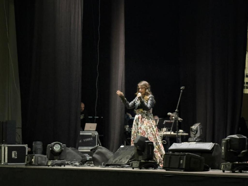 A female singer performing on stage wearing a floral dress and leather jacket, with audio equipment and stage lights in the background.