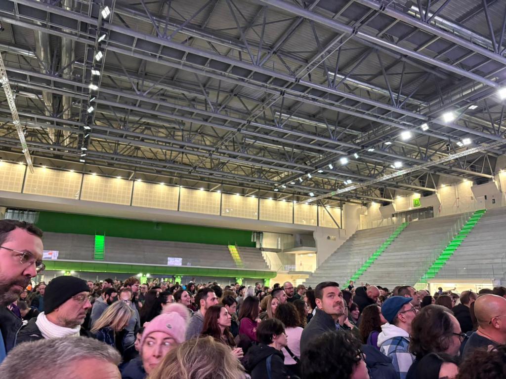 A large crowd of people gathered inside a modern arena, with green lighting and high ceilings, waiting for an event.