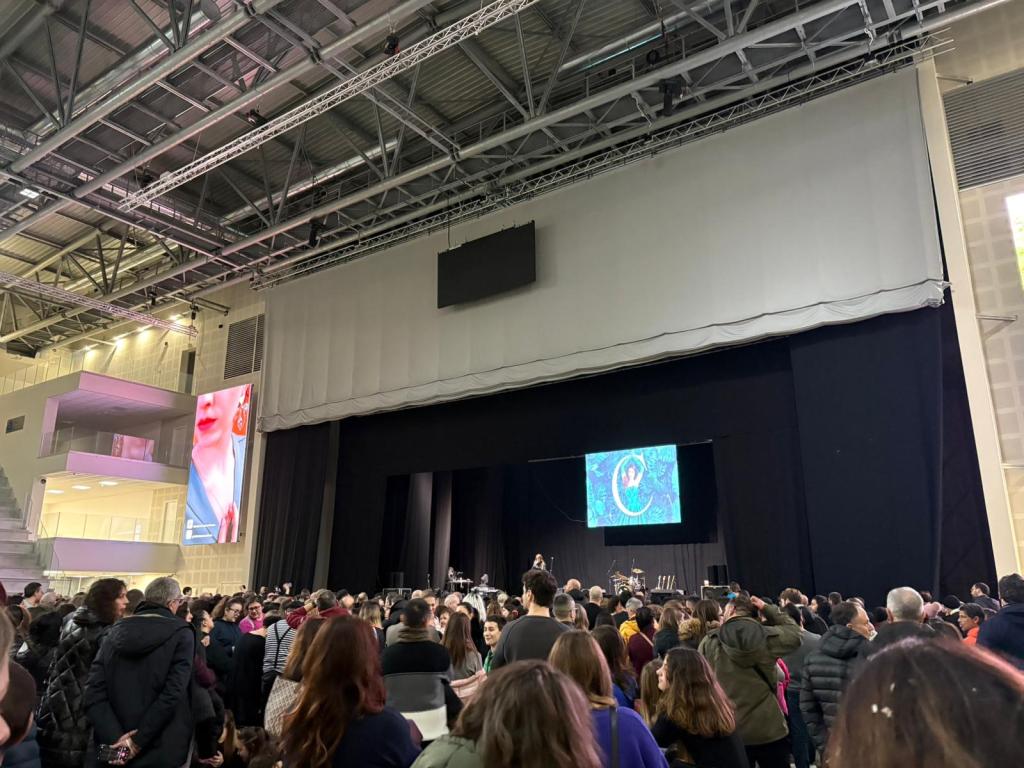 A large crowd gathered in an event hall, with a stage in the foreground. A large screen displays colorful graphics. The venue features high ceilings and modern architecture.