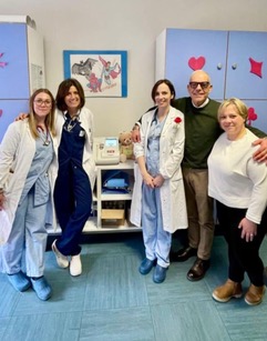 A group of five medical professionals posing for a photo in a brightly colored clinic, wearing white coats and scrubs, with medical supplies in the background.