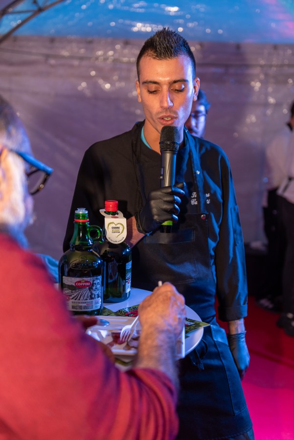 A chef speaking into a microphone while holding a tray with food and bottles of olive oil at a culinary event.