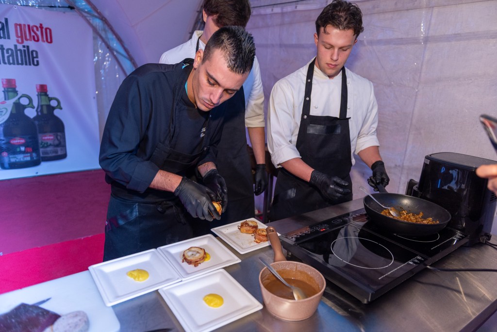 Two chefs preparing dishes at a cooking event, with one chef plating food while the other stirs a pan on the stove.
