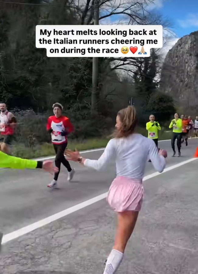 A female runner in a pink skirt looks back while running, smiling at Italian runners cheering her on during a race.