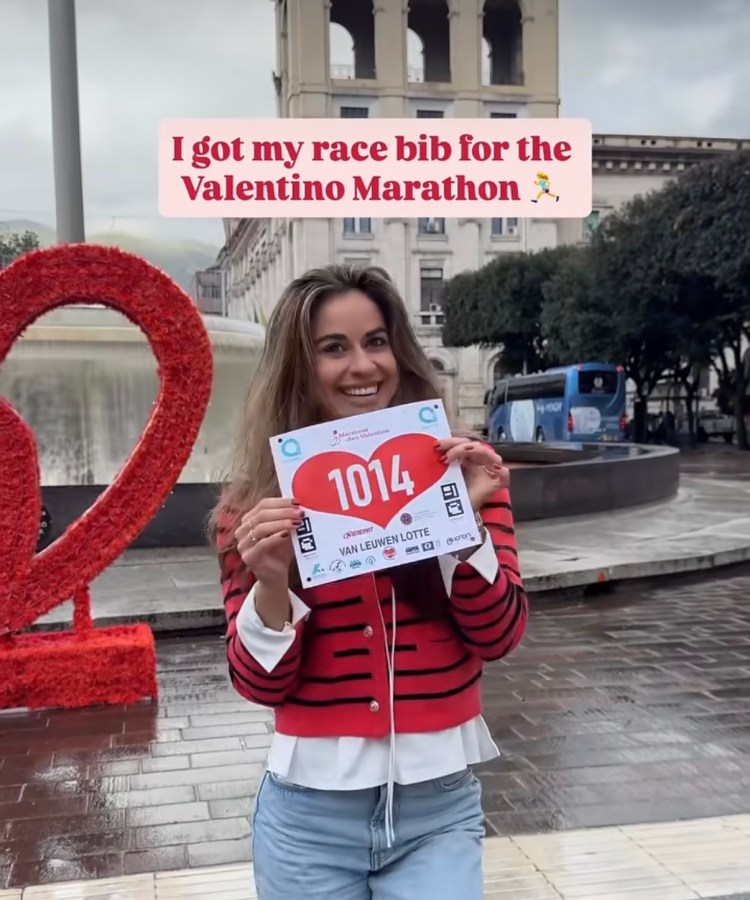 A woman smiles while holding her race bib for the Valentino Marathon, standing in front of a large red '2' sculpture and urban scenery.