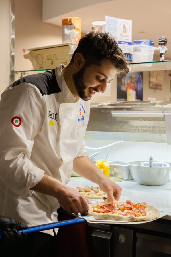 A chef in a white uniform prepares pizza dough with toppings in a kitchen. He is smiling and focused on his task, surrounded by kitchen equipment.
