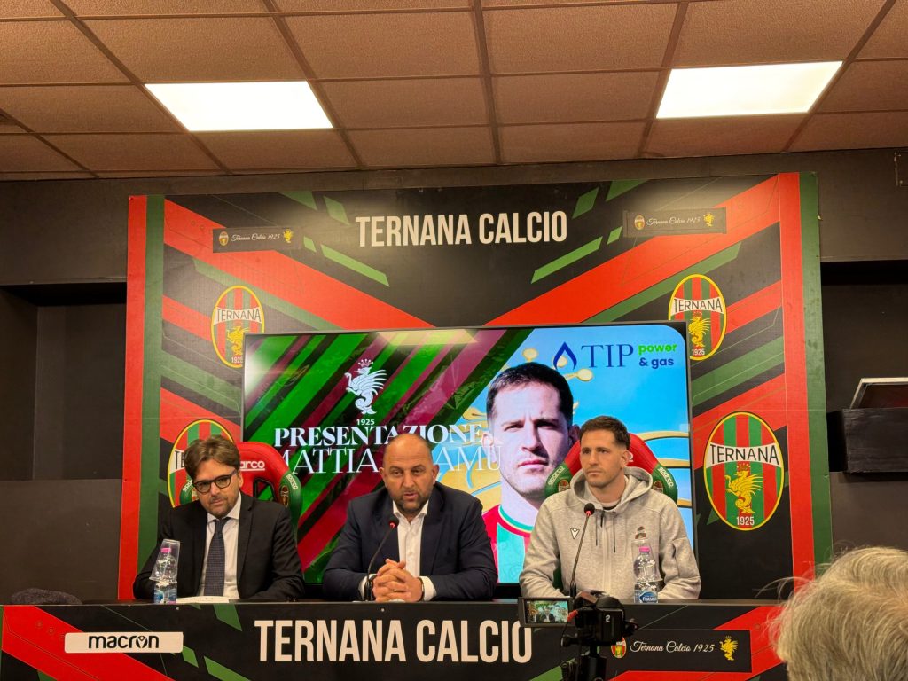 A press conference for the presentation of a new player for Ternana Calcio, featuring three men at a table with a large screen displaying the player's image in the background.