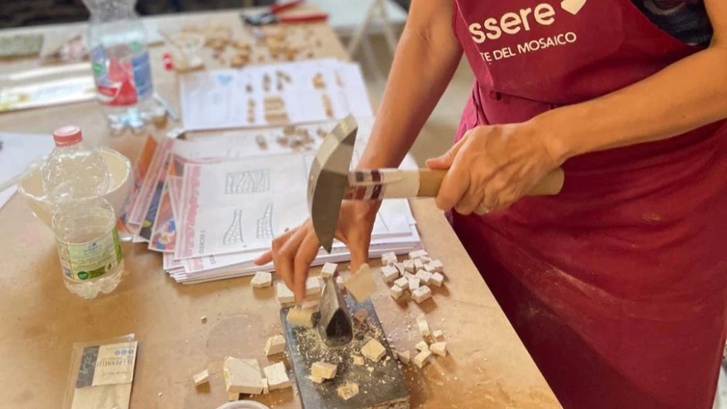 A close-up of a person's hands working on mosaic art, using a hammer to break tiles on a work surface covered with small pieces of stone and papers with mosaic patterns.