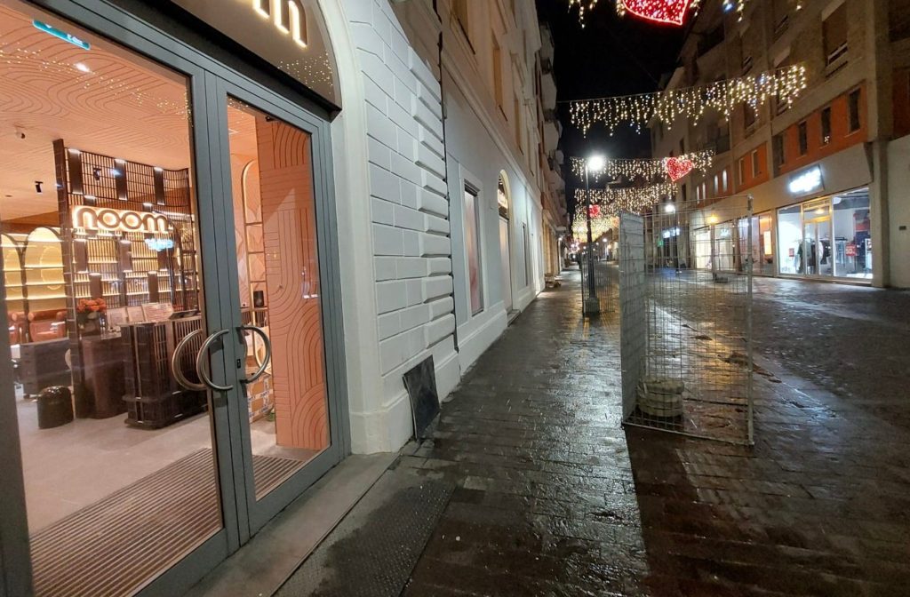 A nighttime street view featuring the entrance of a shop named 'noom' with large glass doors, illuminated by warm lighting. The surrounding area includes festive lights hanging overhead and a cobblestone pavement reflecting the glow. Nearby buildings showcase various storefronts.