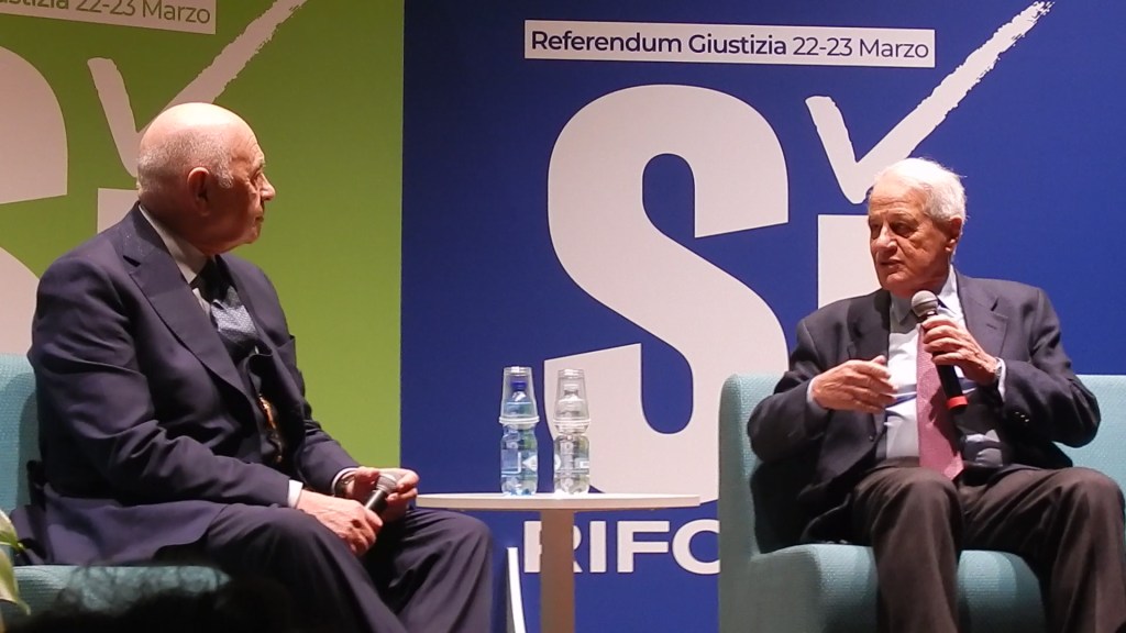 Two men seated on stage during a discussion, with one speaking into a microphone. The backdrop features promotional materials related to a referendum.