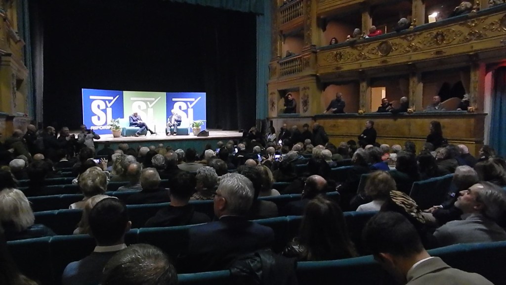 A large audience gathered in a theater for a discussion event, featuring two speakers on stage. The backdrop displays colorful banners. The venue has ornate architecture and an engaged crowd.