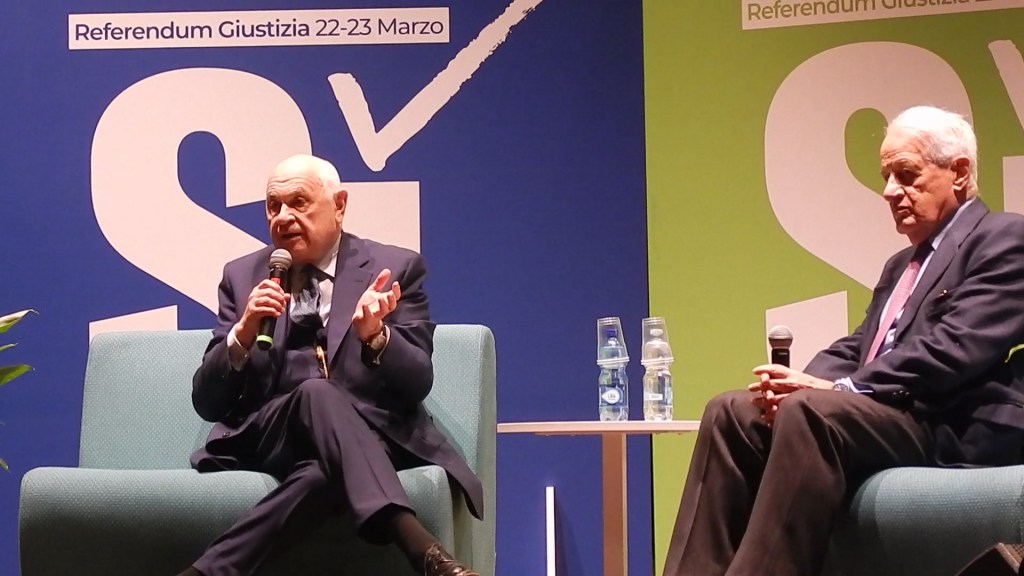 Two men sitting on a stage during a discussion event, with one speaking into a microphone and gesturing, while the other listens attentively. The background features promotional text related to a referendum on justice.