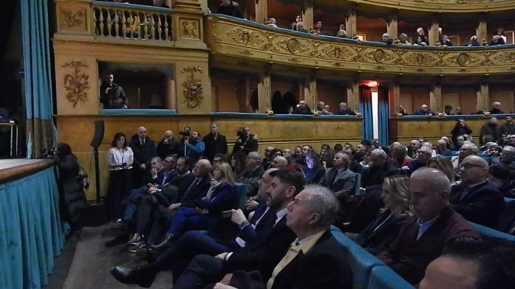 Audience seated in a beautifully decorated theater, listening attentively to a speaker on stage, with some attendees standing and several cameras visible.
