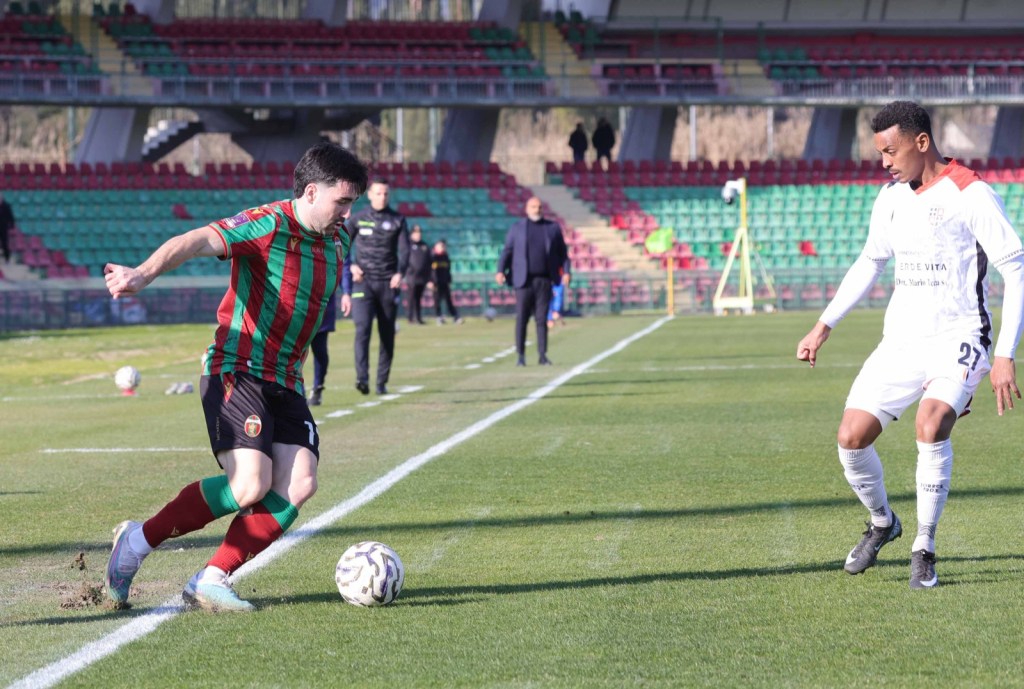A football player in a red and green striped jersey is dribbling a ball on the field, while an opposing player in a white jersey is positioning himself nearby. The scene takes place in a stadium with empty spectator seats in the background.