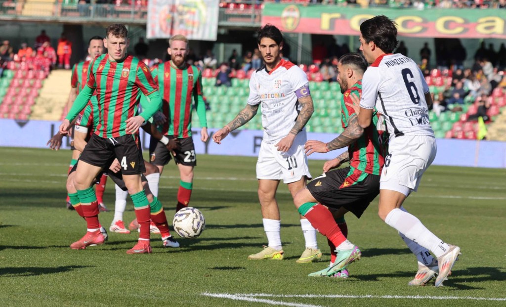 A soccer match in progress with players in red and green striped jerseys competing against those in white jerseys. The ball is on the field as players from both teams engage in play.