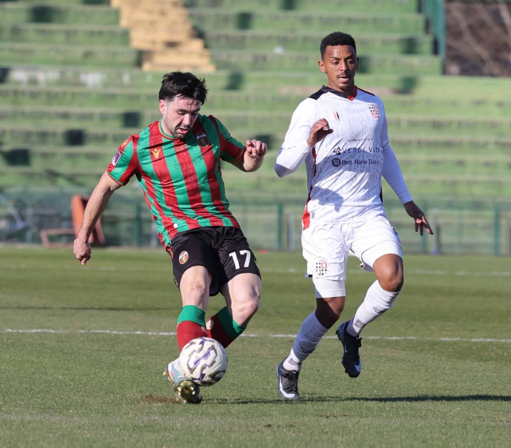 Two soccer players competing for the ball on a grassy field. One player, wearing a red and green striped jersey, is in a dynamic position as he attempts to maneuver the ball, while the other player in a white jersey is closely pursuing.