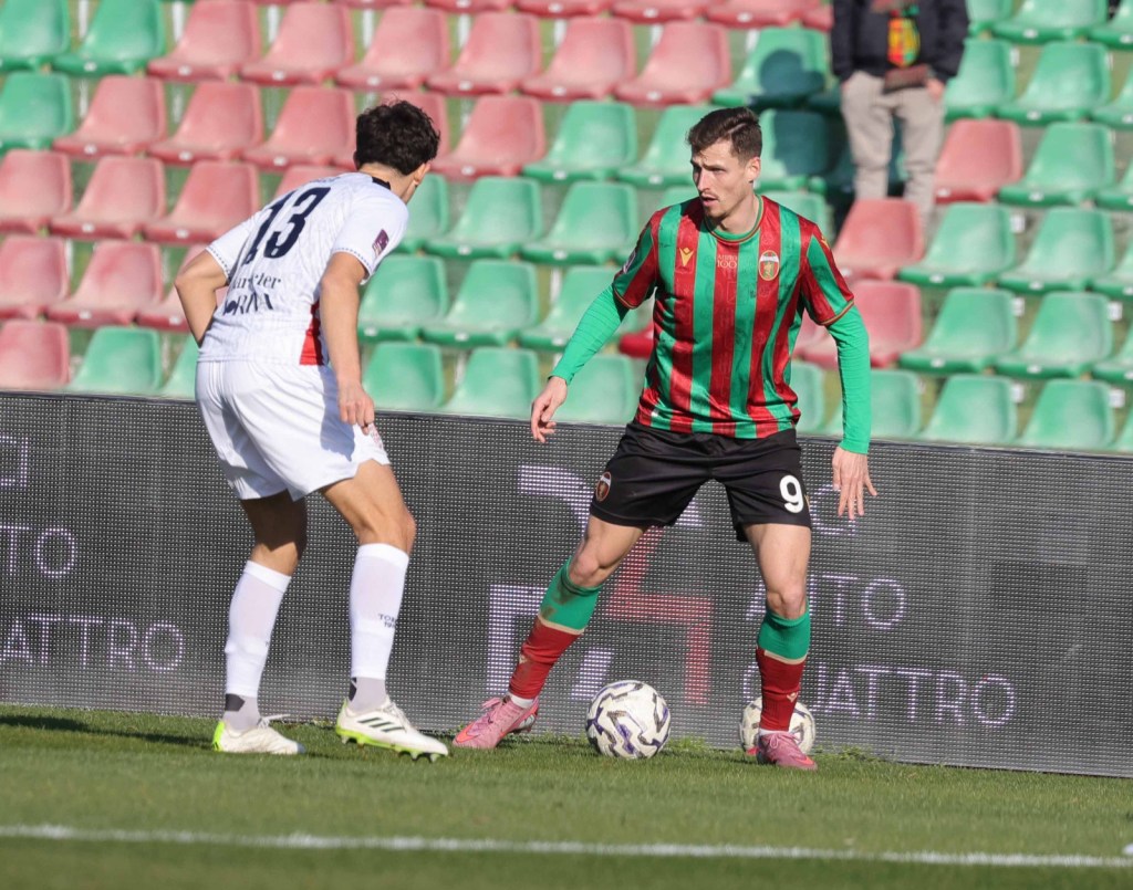 A soccer player in a red and green striped jersey with the number 9 dribbles the ball on the field, while an opposing player in a white jersey with the number 13 positions himself nearby.