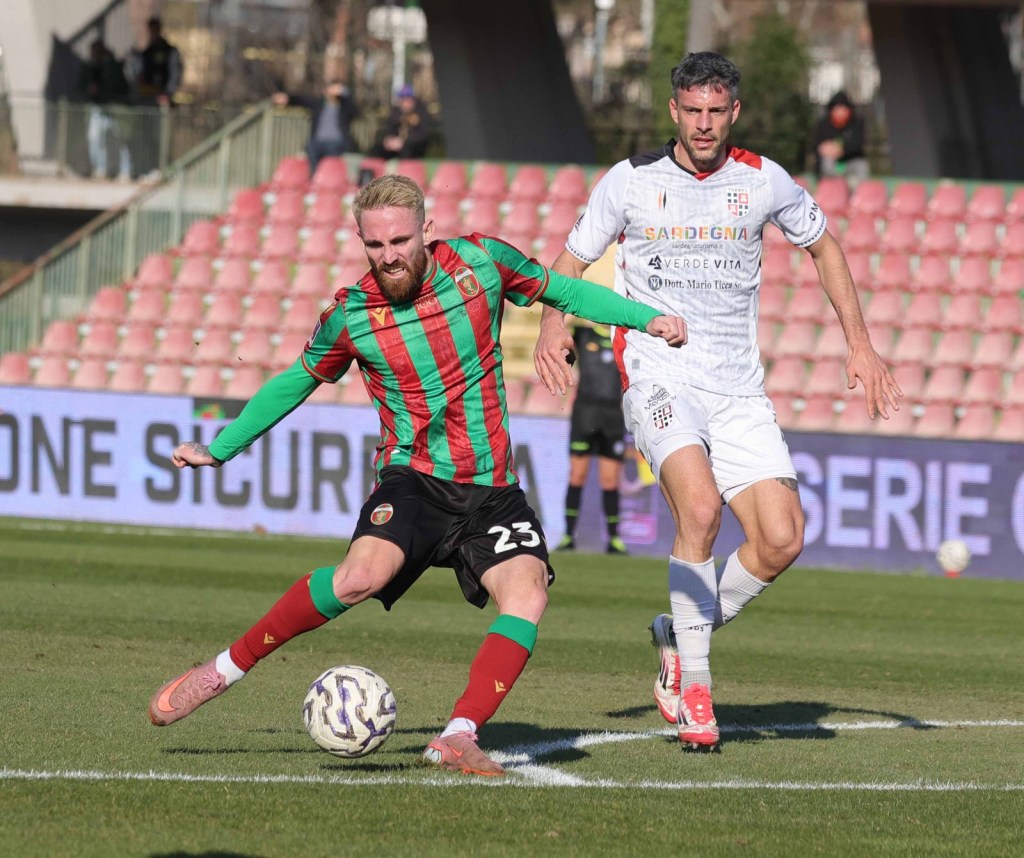 A soccer player in a red and green striped jersey with the number 23 is kicking a soccer ball while another player in a white jersey approaches from behind during a match on a grassy field.