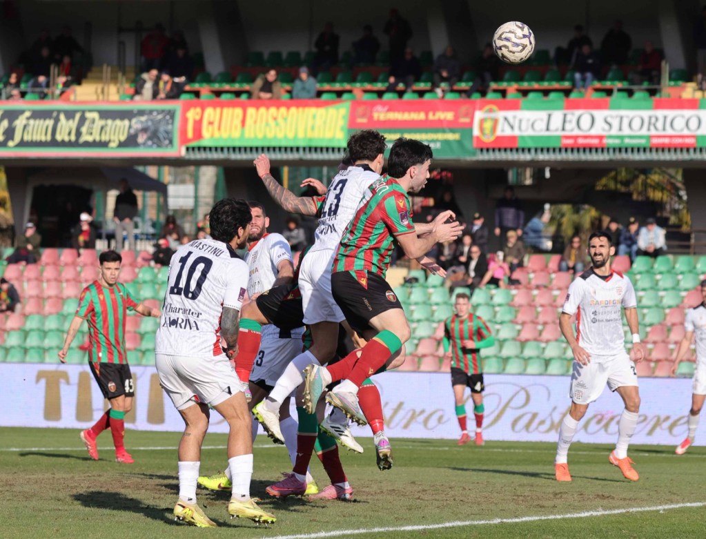 Two soccer players leap for a header while others watch during a match, with a crowd in the background.