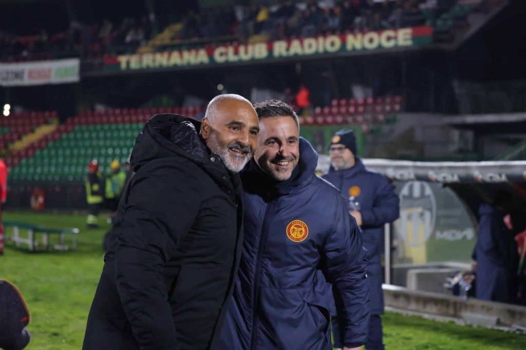 Two smiling men in jackets pose together on a football pitch, with a stadium and fans in the background.