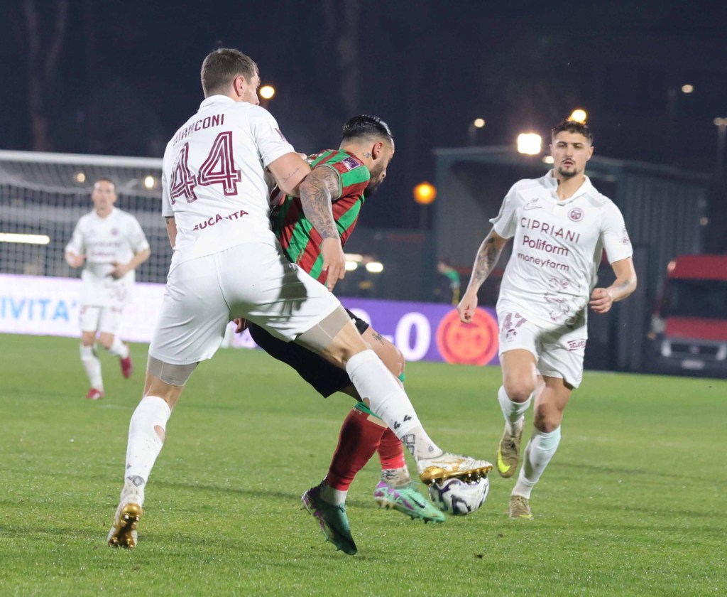 A football match scene with players in action. One player in a red and green striped jersey is being challenged by an opponent in a white jersey, while another player from the same team watches nearby.