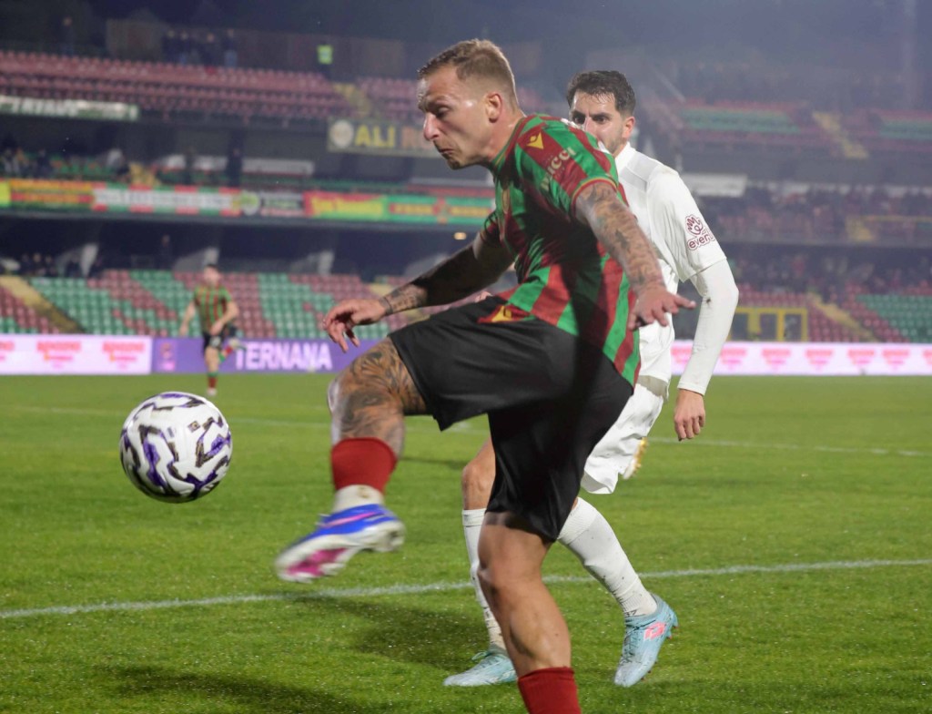 A soccer player in a red and green striped jersey skillfully kicks a ball on the field, with another player in the background watching.