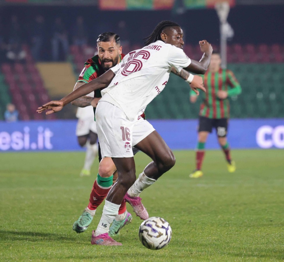 A football match scene showing two players in action, one in a white jersey with the number 16 and the other in a red and green jersey, as they compete for the ball on the pitch.