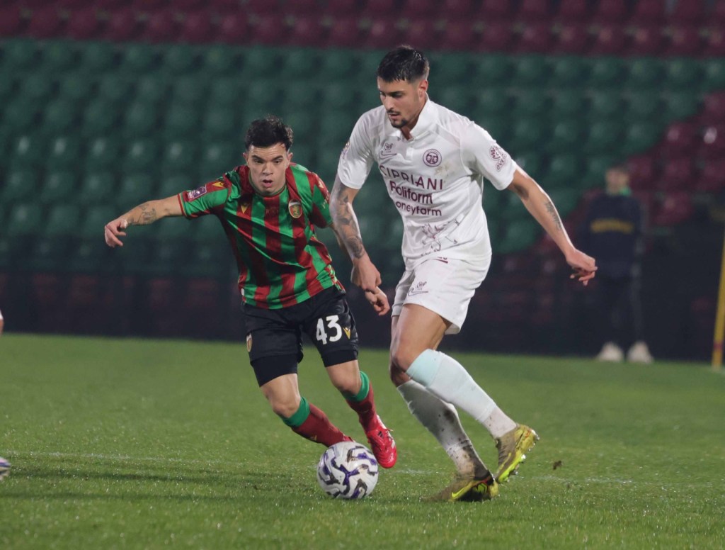 A soccer match scene featuring two players in action; one wearing a green and red striped jersey and the other in a white kit, both vying for the ball on a grass field.