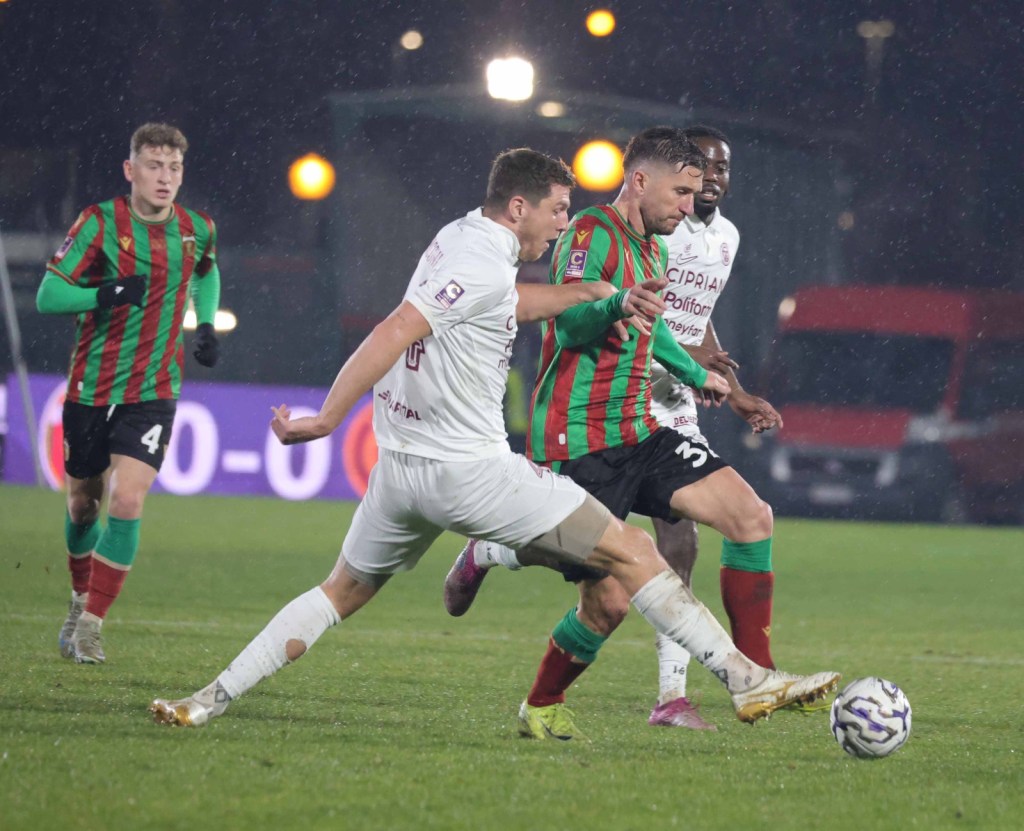 A soccer match in rainy weather, with several players in action. One player in a green and red striped jersey dribbles the ball while being challenged by an opponent in a white jersey. Two other players observe the play from the background.
