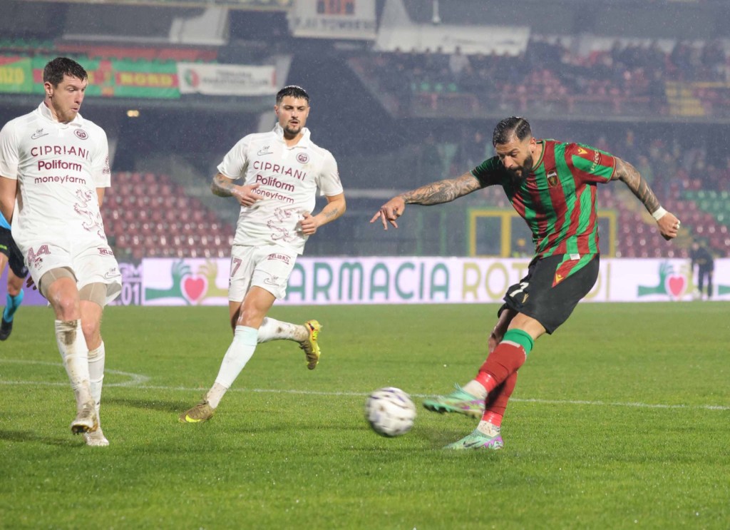 A soccer player in a striped red and green jersey takes a shot on goal while two opposing players in white jerseys attempt to block him. The scene takes place on a wet grass field during a match.