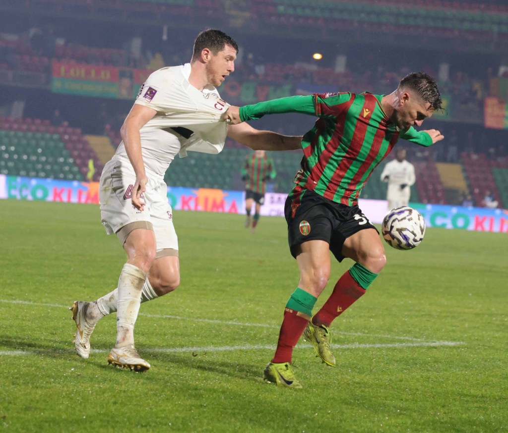 A football match scene showing two players in an intense moment of competition, with one player in a white jersey pulling the shirt of another player wearing a green and red striped jersey.