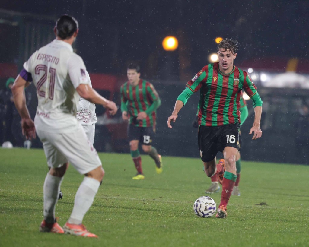 A soccer player in a red and green striped jersey dribbles the ball during a rainy match, with an opposing player in a white jersey and the number 21 in focus.