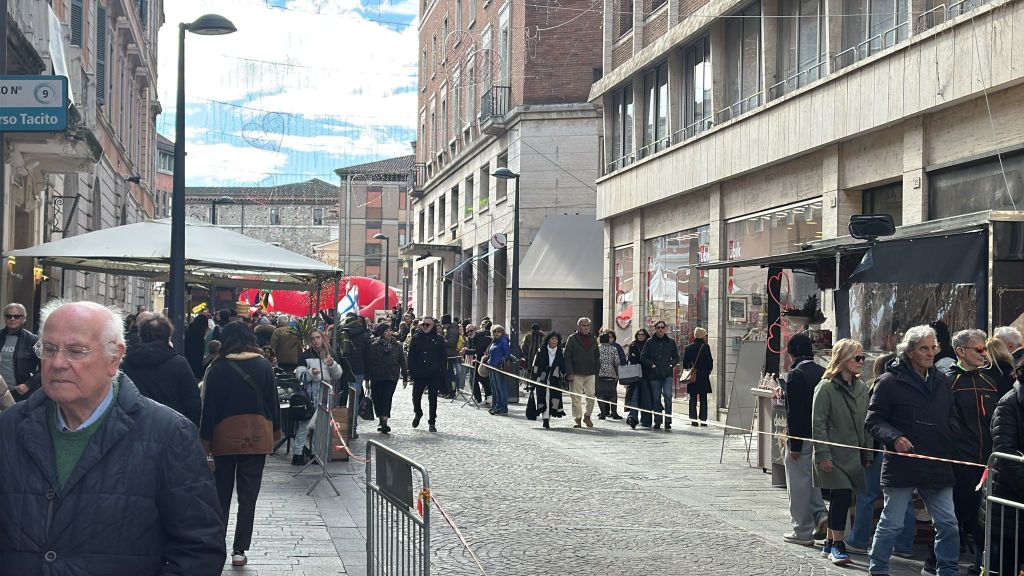 A busy urban street lined with people walking past shops and outdoor seating, with a clear blue sky and fluffy clouds above.