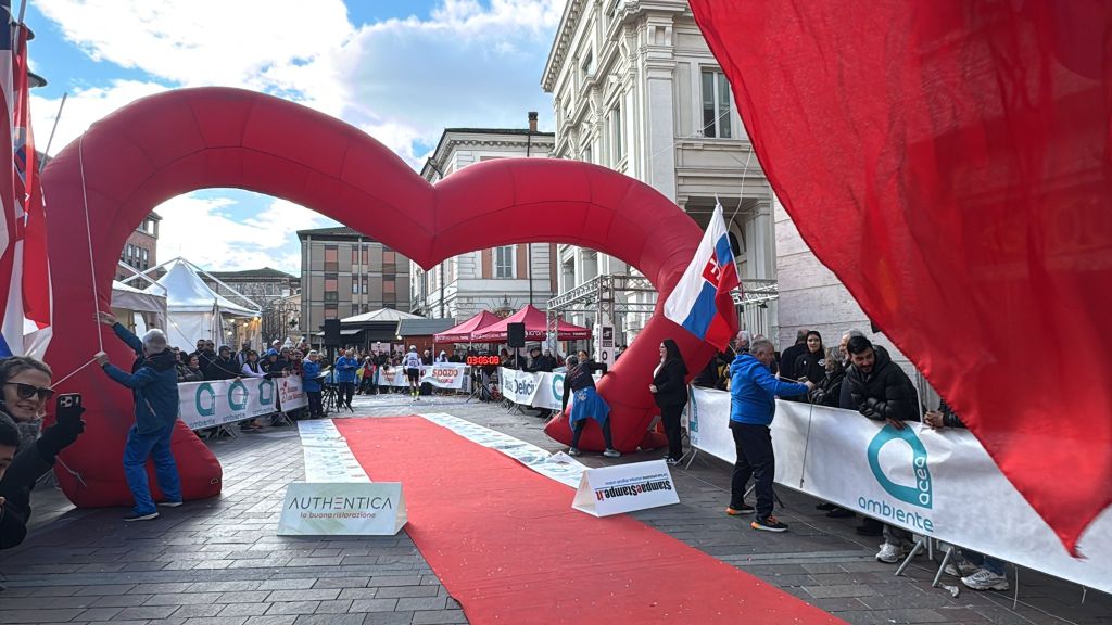 An outdoor event scene with a large red heart-shaped archway and a red carpet. People gather around the arch, participating in what appears to be a race or festival, with tents and banners in the background.