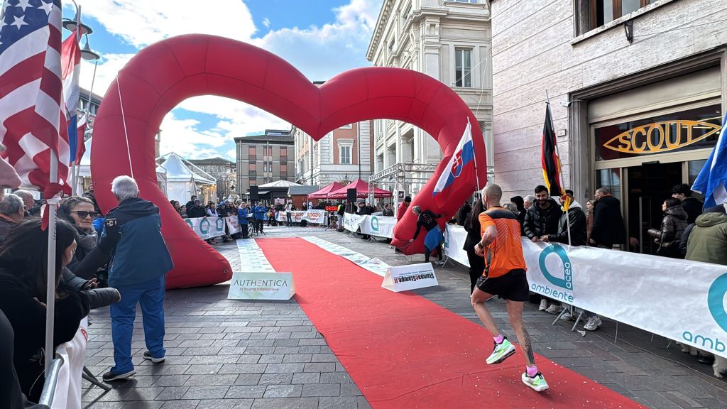 Runner crossing the finish line under a large red heart-shaped arch at a race event, with spectators and flags in the background.