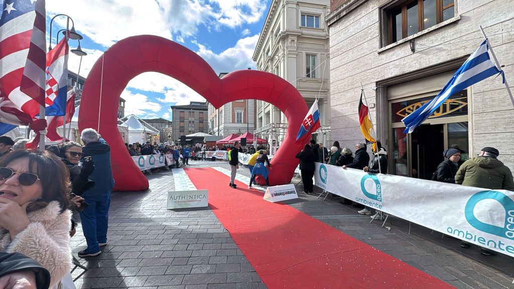 A large red heart-shaped arch marks the entrance to an event on a cobblestone street, with flags and banners in the background. A crowd of people is gathered along the sides of a red carpet leading through the arch.