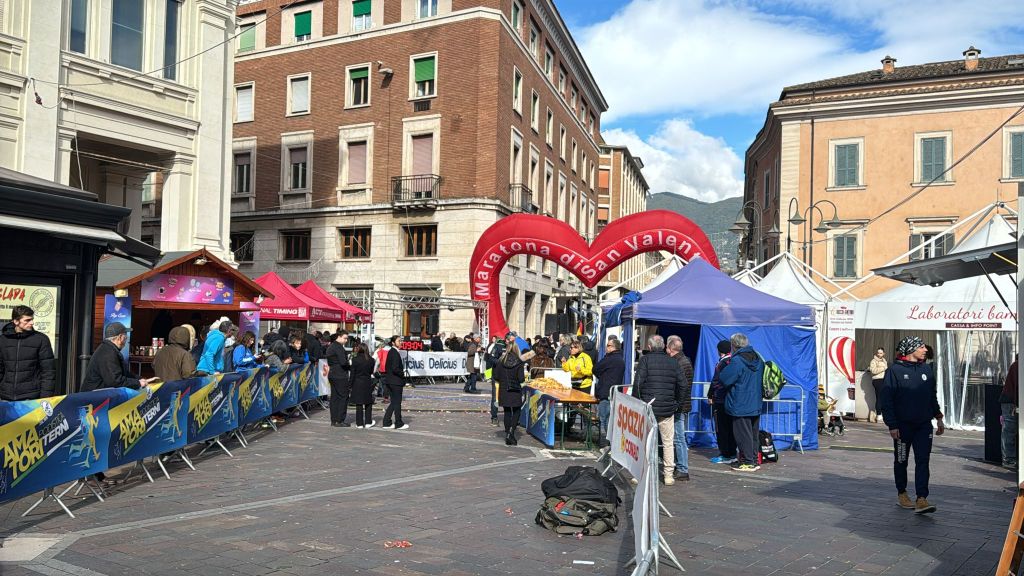Outdoor event scene with festive decorations, including a red heart-shaped arch that reads 'Maratona di San Valentino', colorful tents, and a crowd of people in a busy street.