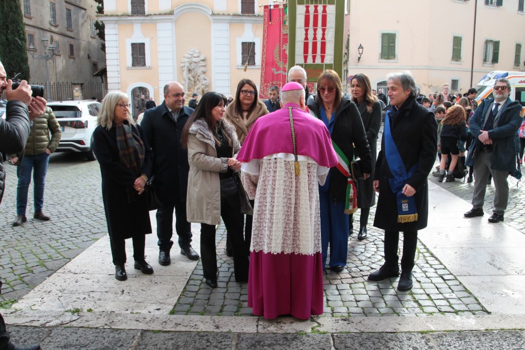 A group of people, including a dressed cleric, interacting in a plaza with historical buildings in the background. Some individuals are wearing formal attire, and others are taking photographs. A colorful banner is visible in the scene.