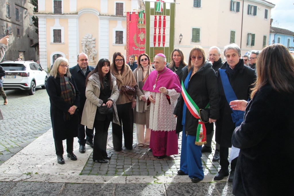 A group of people gathered outdoors in a cobblestone square, with a religious figure in traditional attire at the center, surrounded by local officials and community members.