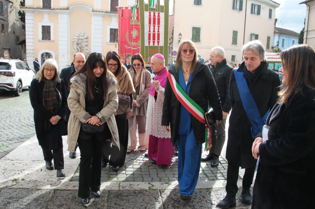 A group of people walking together in a public square, with some wearing formal attire. The scene includes a woman in a mayoral sash, and there are buildings and banners in the background.