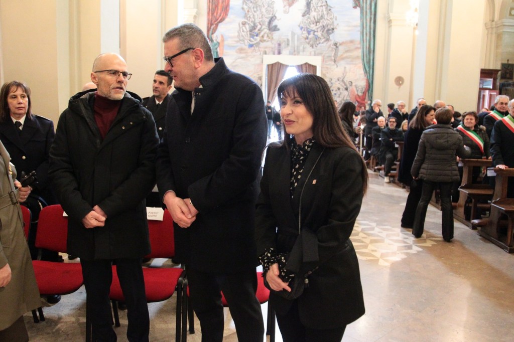 A group of people engaged in conversation inside a hall, with a large mural in the background, and attendees seated in red chairs.