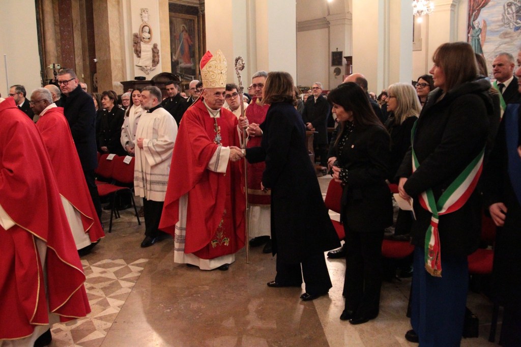 A religious ceremony taking place in a church, featuring a cardinal in red vestments greeting attendees, with people dressed in formal clothing standing around.