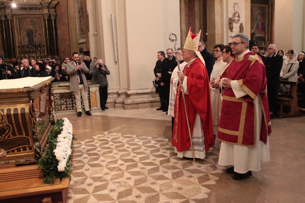 A group of clergy members, including a bishop in red and gold vestments, stand in front of a decorative casket adorned with flowers. The scene takes place in a church, filled with onlookers and cameras capturing the moment.