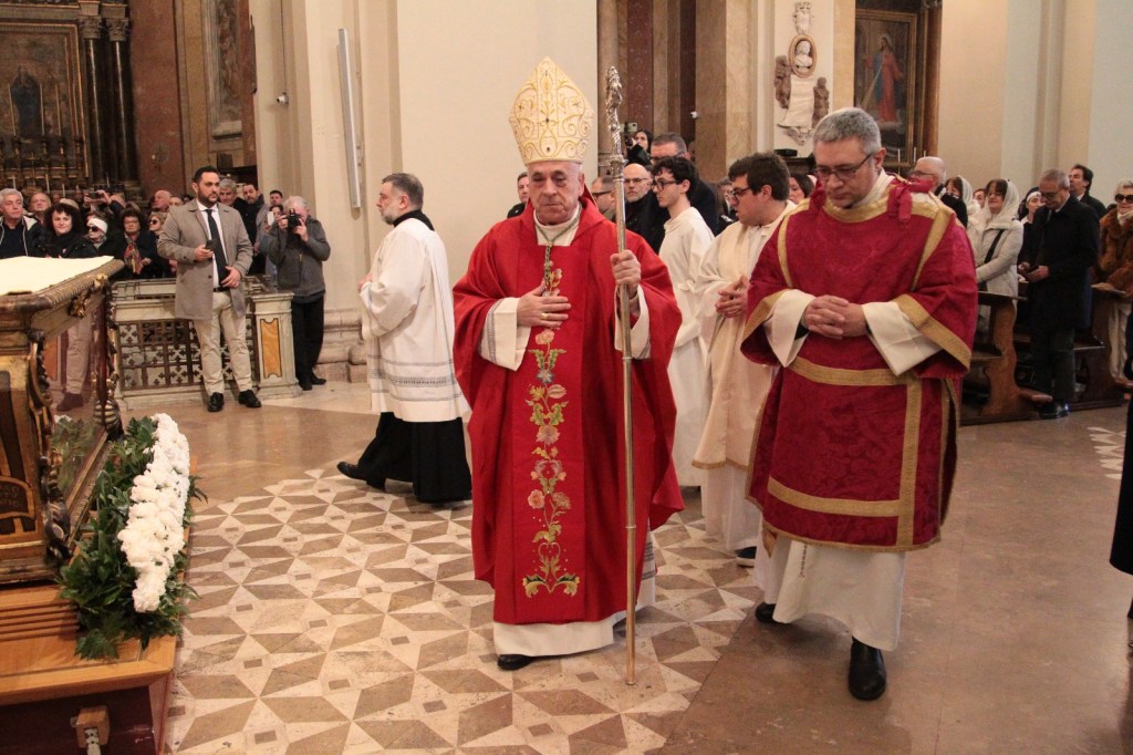 A group of clergy, including a bishop in red vestments and a mitre, process through a church filled with people. The bishop carries a staff, and there are floral decorations nearby.
