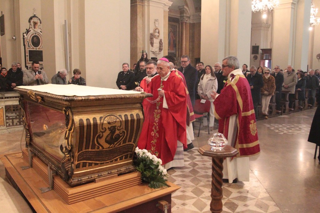 A religious ceremony taking place inside a church, featuring a wooden sarcophagus adorned with golden designs. Clergymen in red vestments are present, along with a congregation of people observing the event.