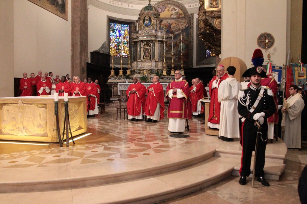 A group of clergy in red robes participates in a religious ceremony inside a church, with a detailed altar and stained glass windows in the background.