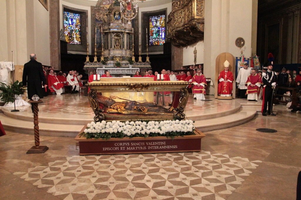 An interior view of a church featuring a ceremonial event. In the foreground, a decorative altar displays a relic encased in glass, surrounded by flowers. Numerous clergy members in red robes are seated in the background, with a cardinal or bishop prominently positioned. Stained glass windows brighten the scene.