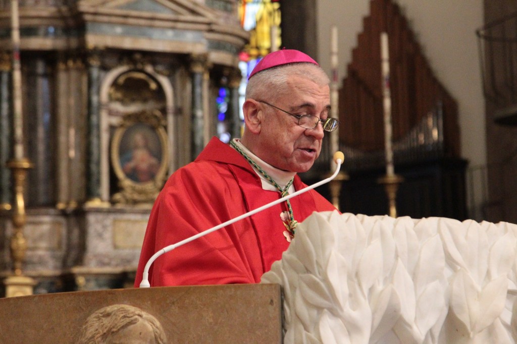 A clergy member wearing a red vestment and a pink mitre delivers a sermon at a church, with ornate architecture and stained glass in the background.