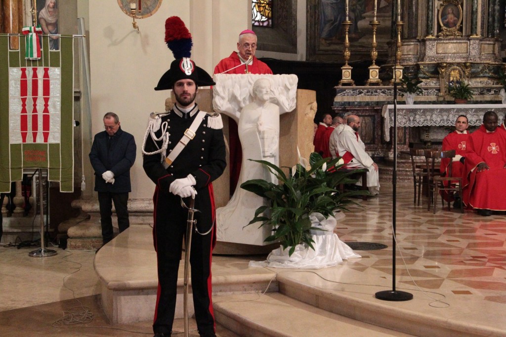 A formal ceremony inside a church, featuring a soldier in historical uniform standing guard, a priest delivering a speech, and attendees in religious attire.