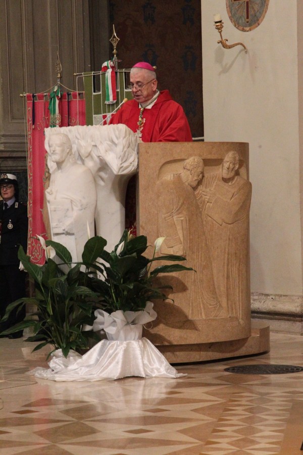 A cardinal in a red robe delivers a speech at a ceremonial event, standing behind a stone podium adorned with sculptures. Green plants are arranged in front of the podium, with a decorated wall in the background.
