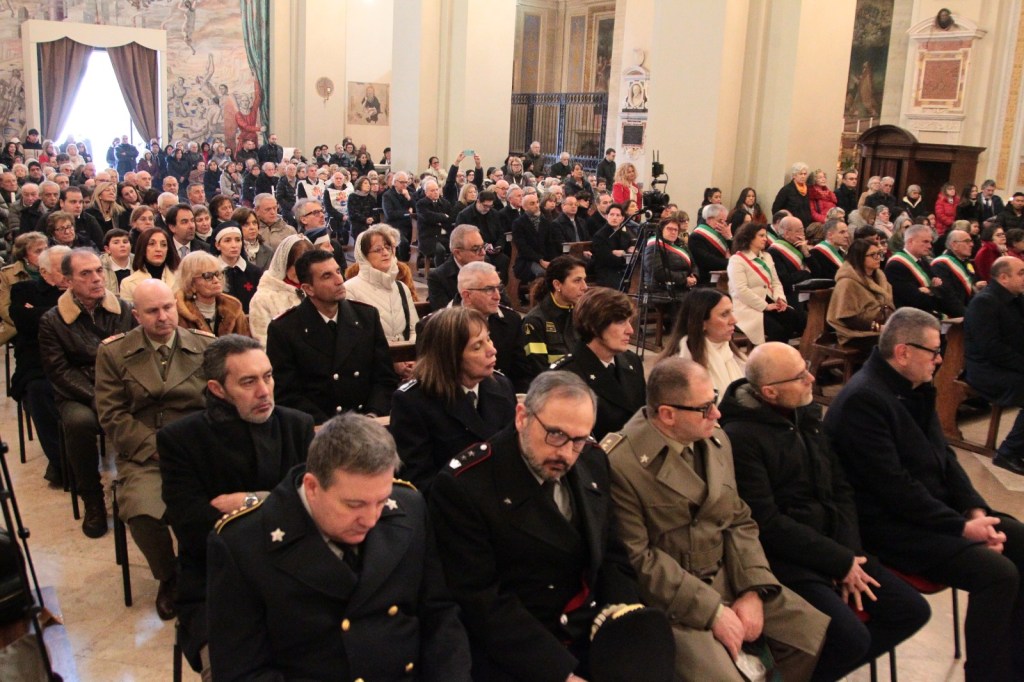 A large audience gathered inside a historical building, seated in rows facing the front. Various attendees, including uniformed officials and civilians, are engaged in the event. The room features elegant architecture and murals.